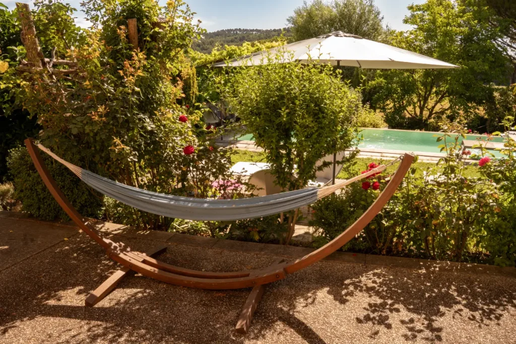 Hängematte in einem Holzgestell vor Büschen, mit Blick auf den Pool.