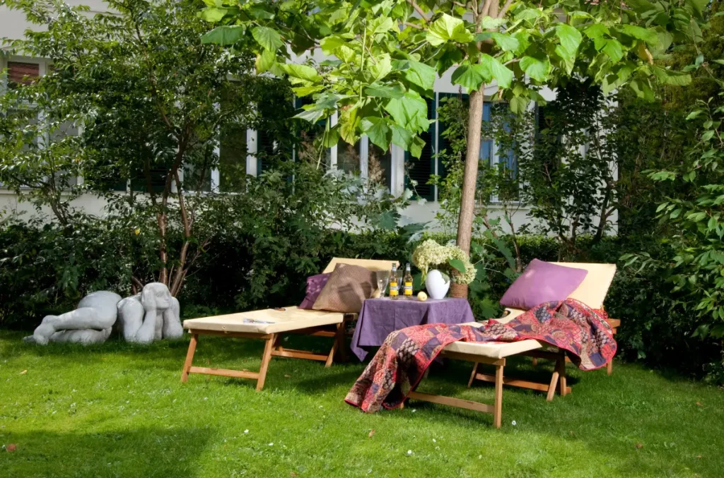 Zwei Holzliegen mit Auflagen, Decke und Kissen unter einem Baum im Garten, dazwischen ein kleiner Tisch mit Getränken.