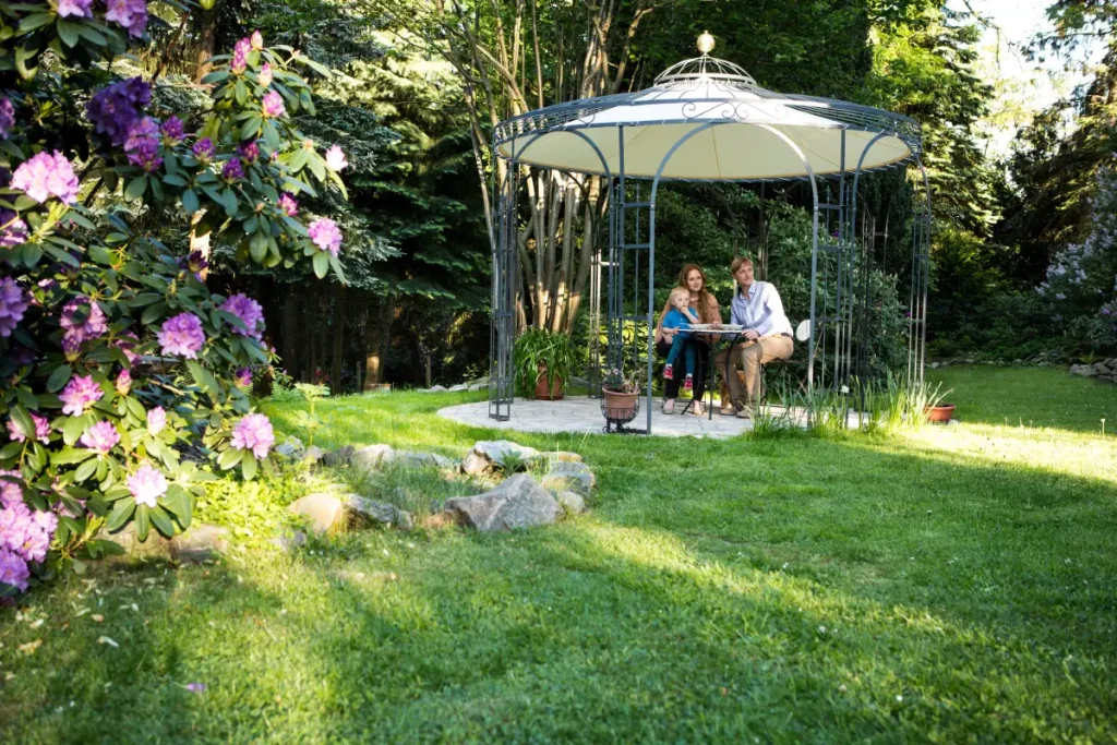 Romantisch runder Metallpavillon auf gepflastertet Fläche, umgeben von gepflegtem Rasen, Bäumen und Büschen.