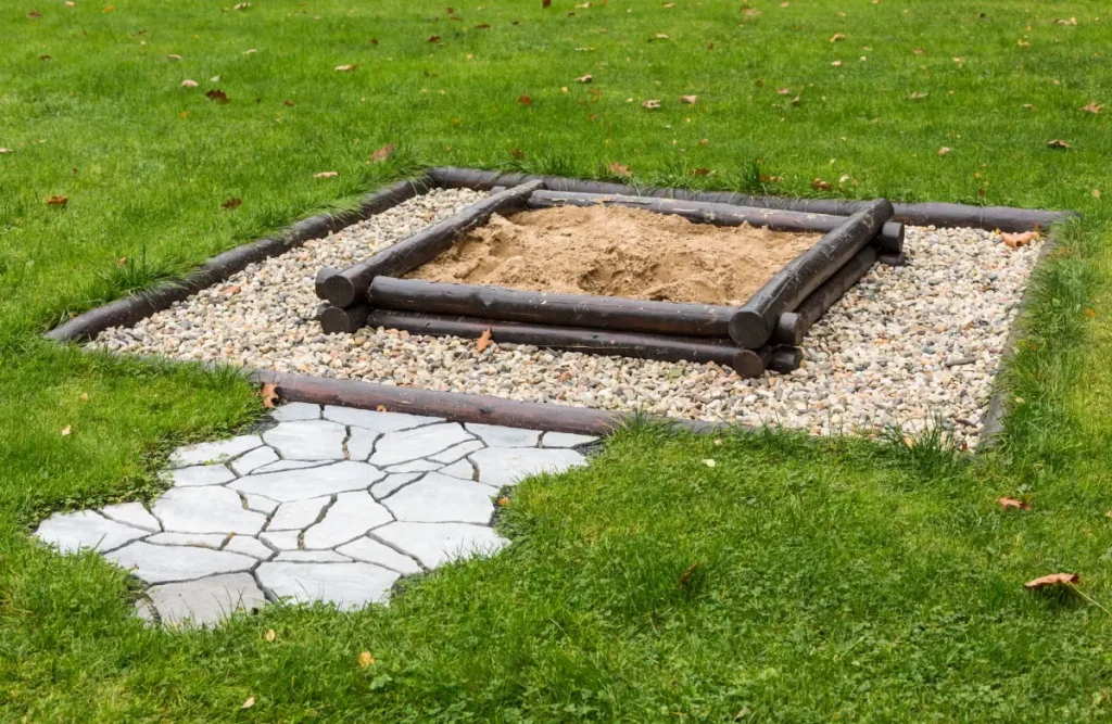 Sandkasten mit edler Umrandung aus Rundhölzern, umgeben von groben Steinen, eingefasst durch weitere Rundhölzer. Ein kurzer Weg aus Natursteinen lädt zum Spielen ein.