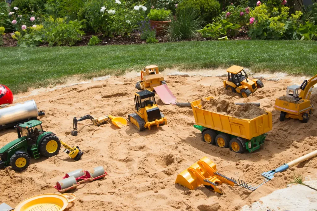 Ebenerdiger Sandkasten, umrandet mit Steinen, gefüllt mit Traktor, Bagger und Hänger.