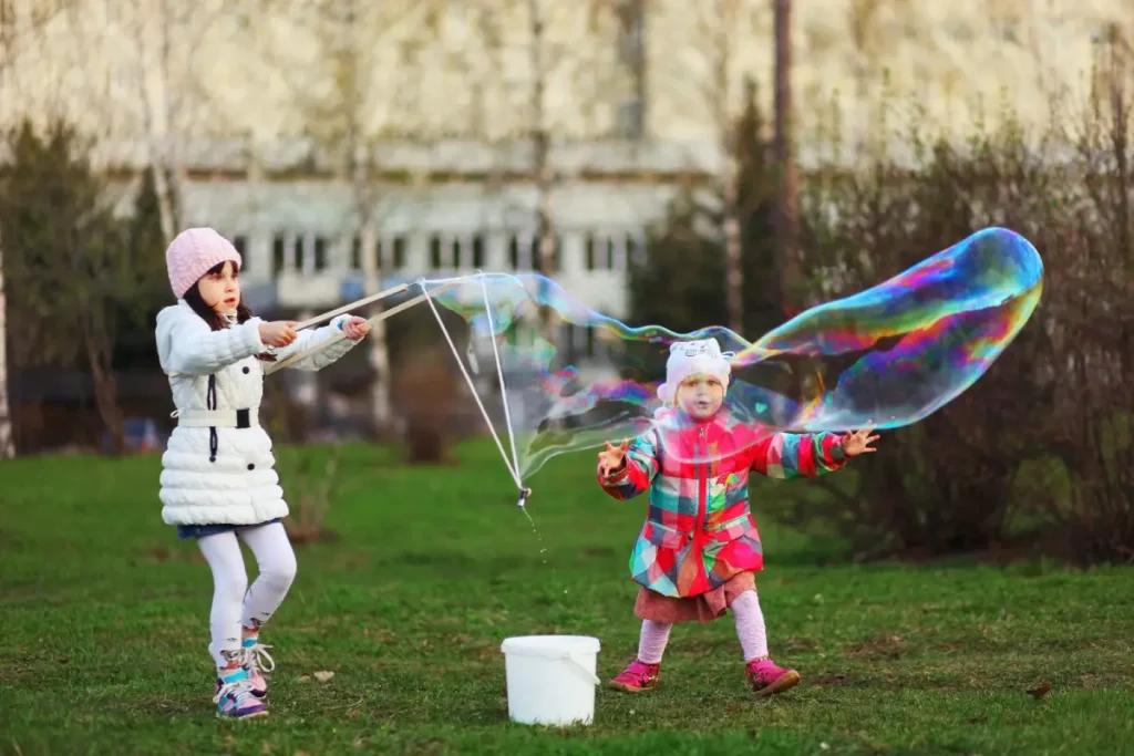 Mädchen macht mit ihrer kleinen Schwester riesige Seifenblasen im Garten Gartenspielzeug: 8 einfache DIY-Ideen für Ihre Kinder